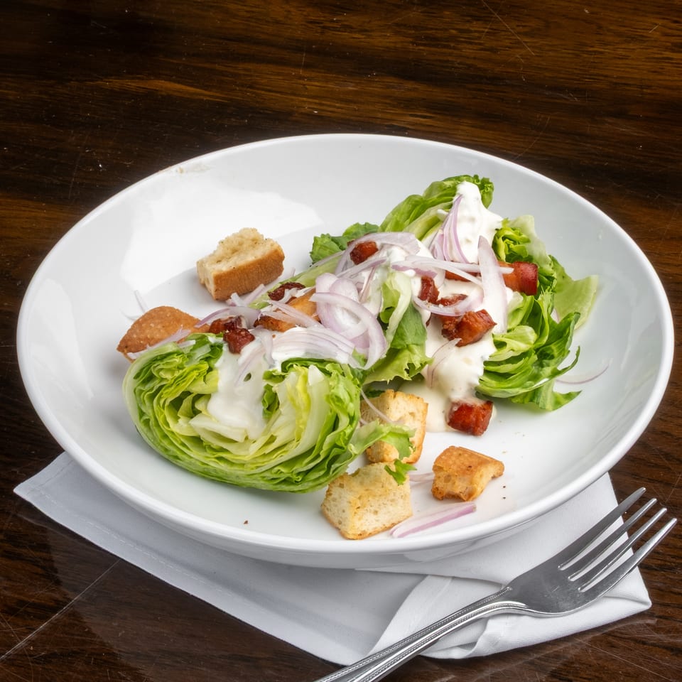 Wedge salad with lettuce, bacon, red onion, croutons, and creamy dressing in a white bowl.