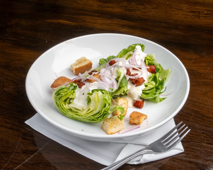 Wedge salad with lettuce, bacon, red onion, croutons, and creamy dressing in a white bowl.
