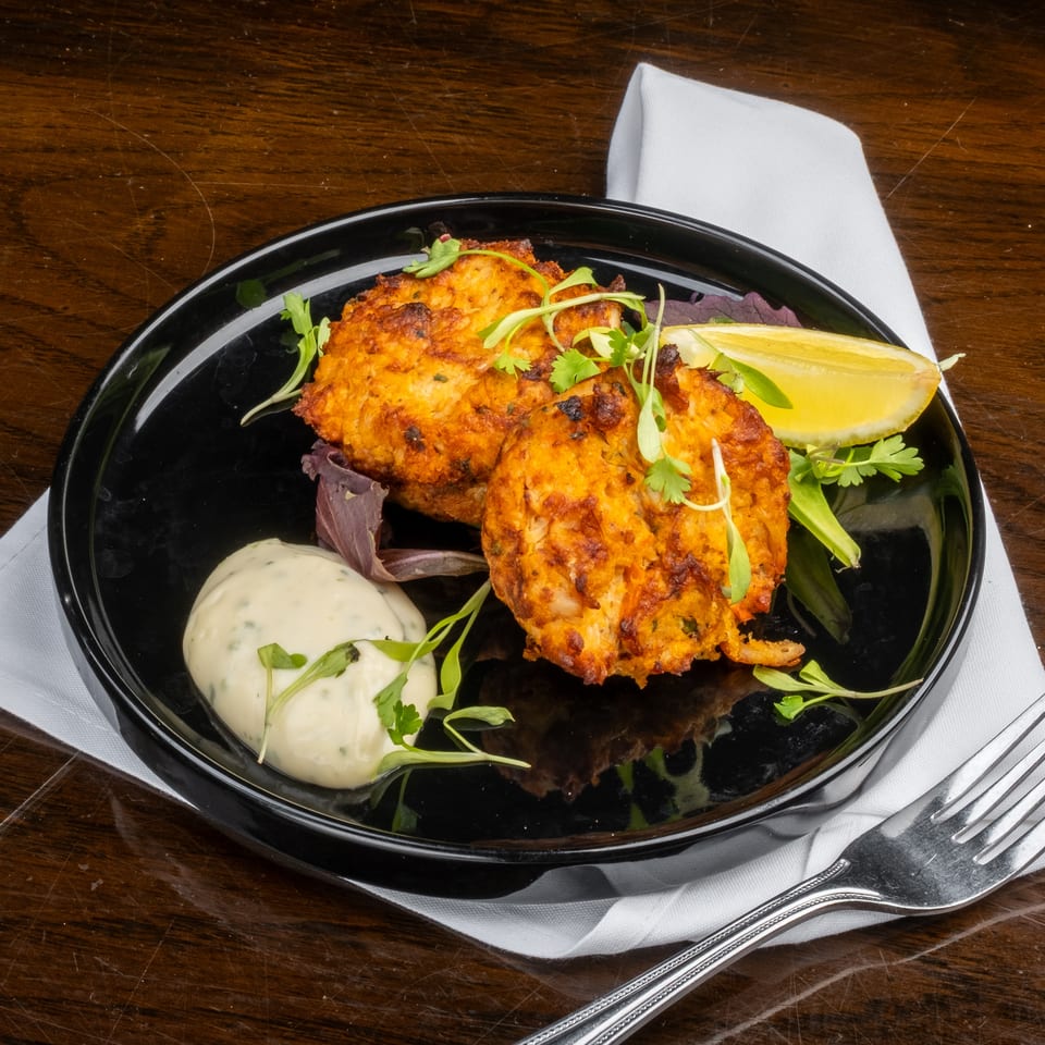 Two golden-brown crab cakes with a lemon wedge, sauce, and greens on a black plate.