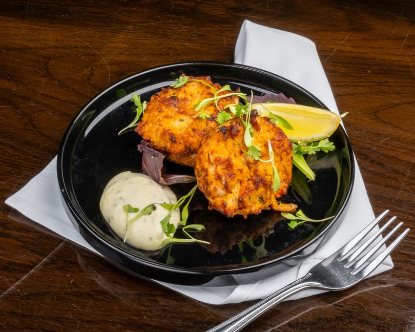 Two golden-brown crab cakes with a lemon wedge, sauce, and greens on a black plate.