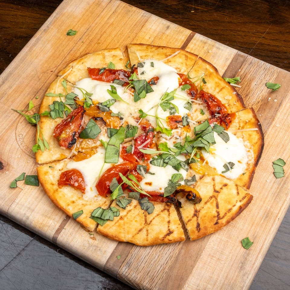 Grilled flatbread pizza with melted mozzarella, roasted tomatoes, and fresh basil on a wooden board.