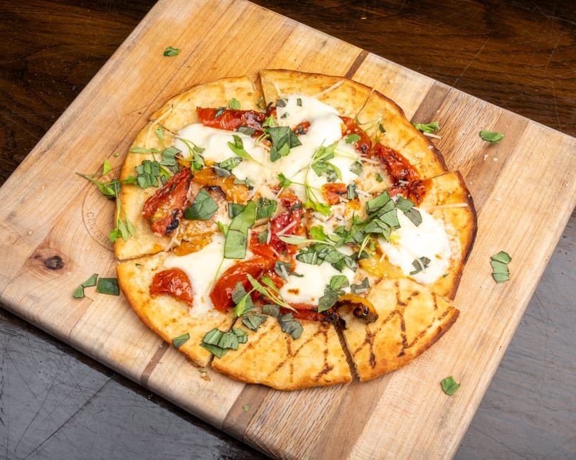 Grilled flatbread pizza with melted mozzarella, roasted tomatoes, and fresh basil on a wooden board.