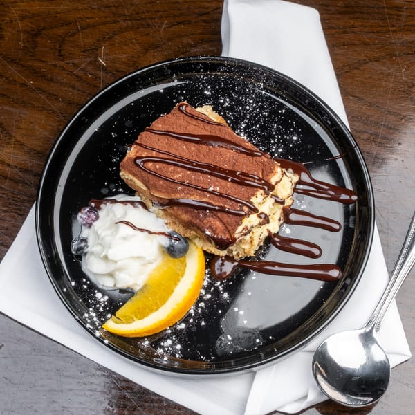 Tiramisu with chocolate drizzle, whipped cream, blueberries, and an orange slice on a black plate.