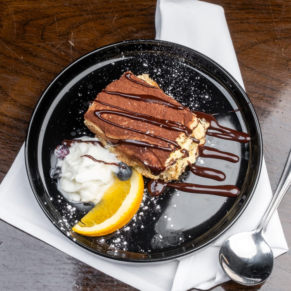 Tiramisu with chocolate drizzle, whipped cream, blueberries, and an orange slice on a black plate.