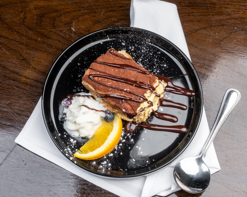 Tiramisu with chocolate drizzle, whipped cream, blueberries, and an orange slice on a black plate.