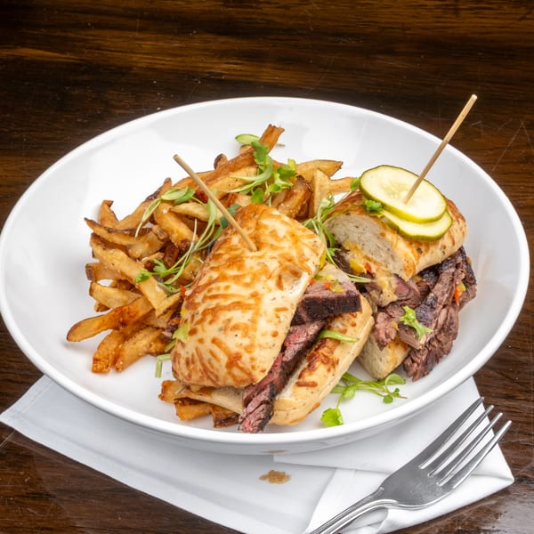 Steak sandwich with melted cheese, pickles, and fries, garnished with greens, on a white plate.
