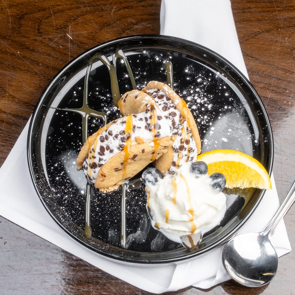 Two chocolate chip cookie tacos filled with ice cream, drizzled with caramel, on a black plate.