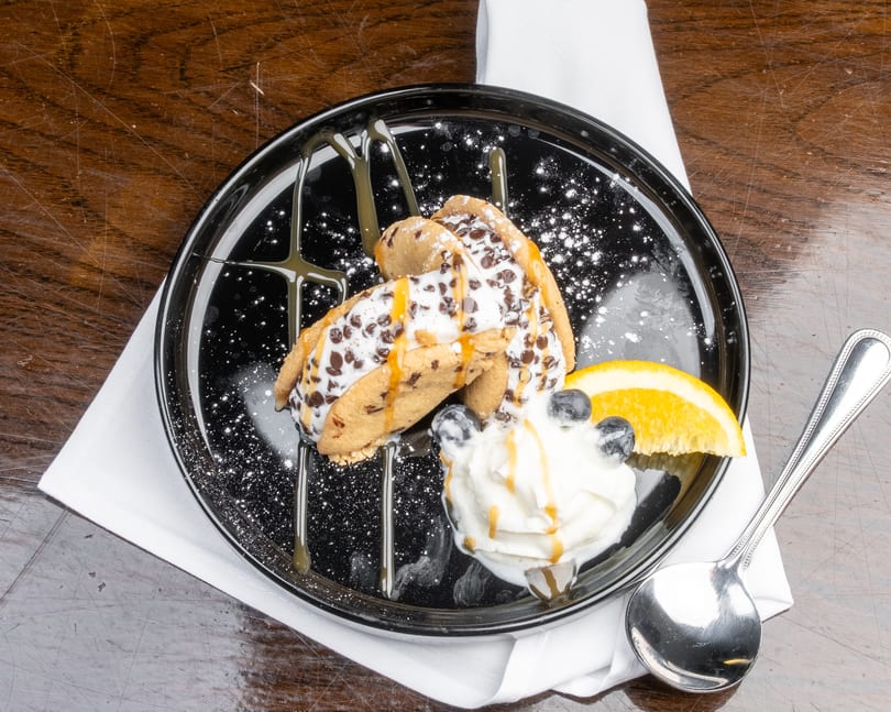 Two chocolate chip cookie tacos filled with ice cream, drizzled with caramel, on a black plate.