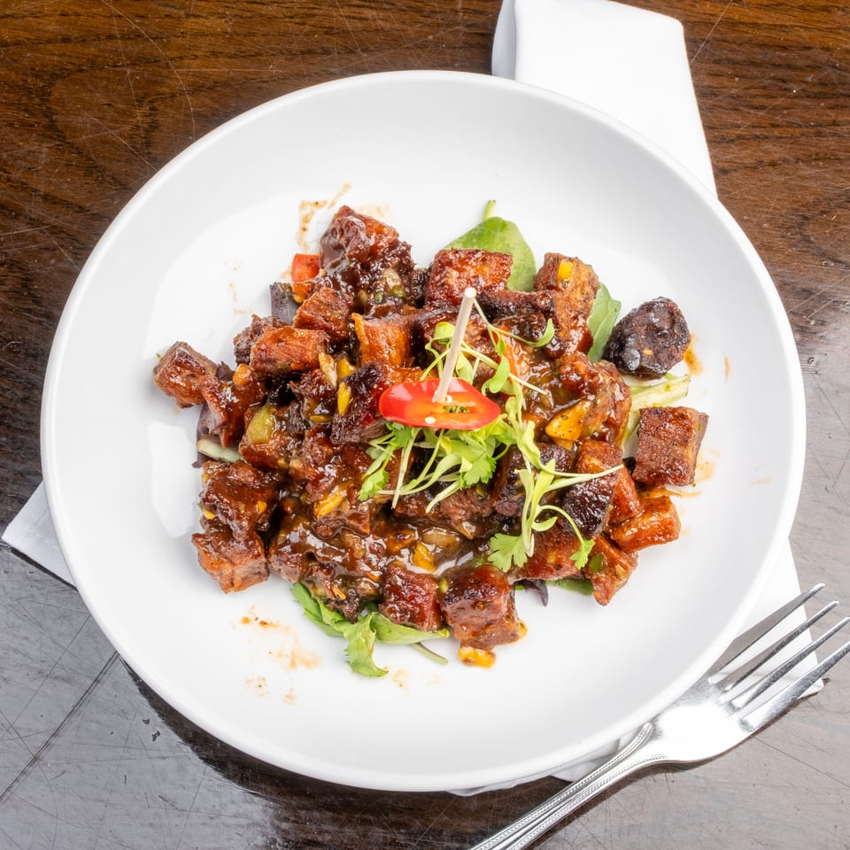 A white plate filled with glazed pork belly cubes, garnished with microgreens and a red chili.