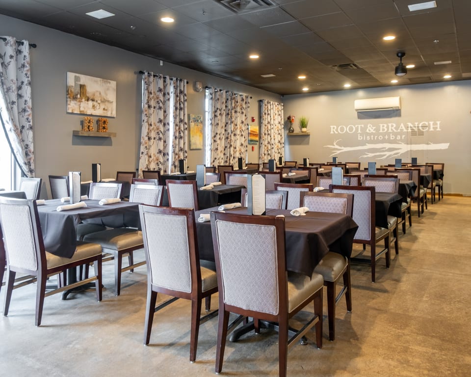 Interior of Root & Branch Bistro + Bar, showing rows of dining tables with dark tablecloths and upholstered chairs.